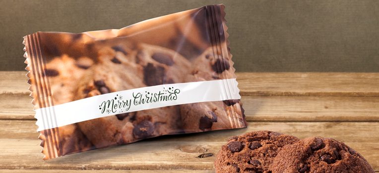 Un paquet de biscuits aux pépites de chocolat portant l'inscription « Joyeux Noël » est posé sur une surface en bois. Deux biscuits aux pépites de chocolat sont disposés à proximité, créant une ambiance festive et rustique.