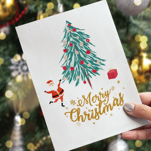 Une main tient une carte de Noël festive représentant un sapin décoré, le Père Noël et un coffret cadeau. La carte porte l'inscription « Joyeux Noël » en lettres dorées. Un sapin de Noël flou et illuminé apparaît en arrière-plan.