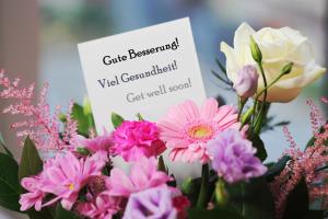 Un bouquet de fleurs roses et violettes, avec une rose blanche. Une carte est glissée au milieu, avec l'inscription « Gute Besserung ! Viel Gesundheit ! Bon rétablissement ! » en allemand et en anglais.