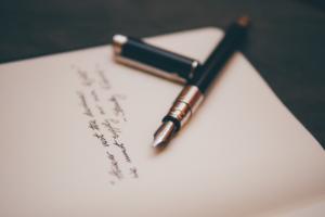 Un stylo-plume, sans son capuchon, repose sur un carnet ouvert. Le papier présente une écriture cursive à l'encre sombre, légèrement floue en arrière-plan. Le stylo et le carnet sont posés sur une surface sombre.