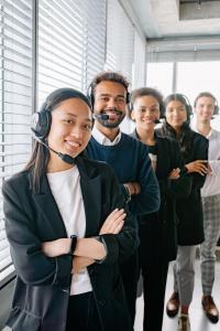 Un groupe hétéroclite de six personnes, portant des casques audio, fait la queue à l'intérieur. Elles sourient, certaines ont les bras croisés. Elles portent une tenue décontractée. En arrière-plan, de grandes fenêtres avec des stores.